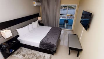 A hotel room with a bed, bedside table with a lamp, a TV mounted on the wall, a small bench, and a view of the water through a sliding glass door.