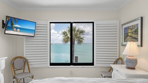 Un dormitorio con un ventanal grande, una cama, un televisor, una silla, una lámpara y un cuadro enmarcado.