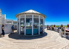 Um gazebo com cobertura de vidro em deck de madeira, cercado por mesas e cadeiras.