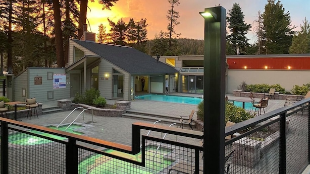 Un espace piscine comprenant un petit bâtiment, une terrasse entourée d’une clôture et une vue sur les arbres et le coucher du soleil.
