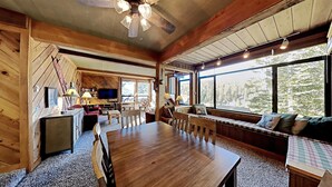 L’intérieur chaleureux d’un chalet avec une table à manger en bois, un canapé recouvert d’un tissu à carreaux et une grande fenêtre donnant sur la forêt.