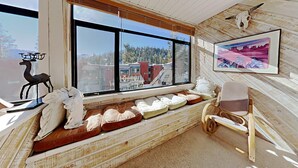 L’intérieur d’un chalet rustique est agrémenté d’une grande fenêtre offrant une vue sur les bâtiments et les arbres. Une sculpture de cerf et un tableau encadré ornent les murs.