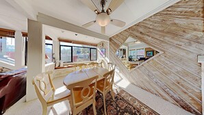 Un espace repas comprenant une grande table et des chaises, un ventilateur de plafond et une vue sur l’extérieur grâce à de grandes fenêtres.
