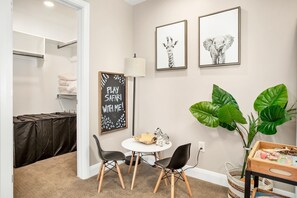 Ein Zimmer mit weißer Wand, einer Tafel mit der Aufschrift 'PLAY SAFARI WITH ME!', zwei gerahmte Tierbilder, ein runder Tisch mit Stühlen, eine große grüne Pflanze und ein Korb mit Spielzeug.