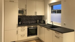 Une cuisine moderne avec des meubles blancs, un plan de travail noir, un four encastré, un four à micro-ondes et une fenêtre avec des stores.