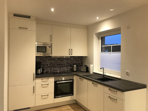 A modern kitchen with white cabinets, a black countertop, a built-in oven, a microwave, and a window with blinds.