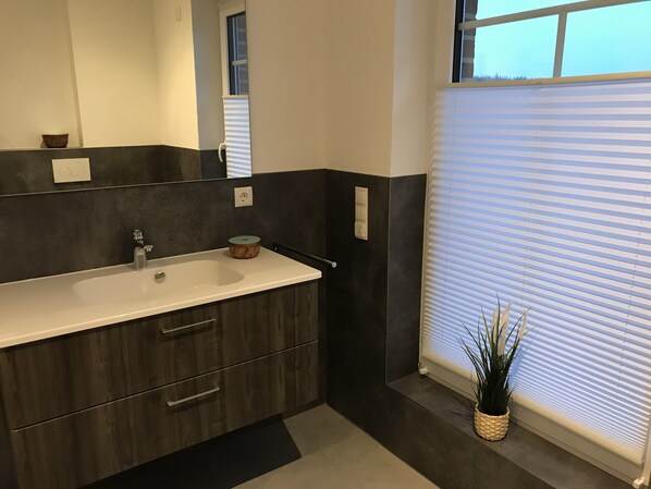 Un baño moderno con un lavamanos grande, muebles de madera oscura, un espejo y una ventana con persianas.