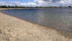 Une plage à l’eau claire, avec un rivage de galets et une ligne d’arbres à l’horizon, sous un ciel partiellement nuageux.