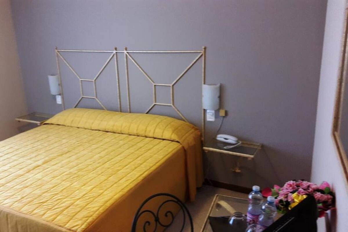 Uma cama com cabeceira geométrica, uma mesinha de cabeceira com um telefone e uma pequena mesa com um vaso de flores.