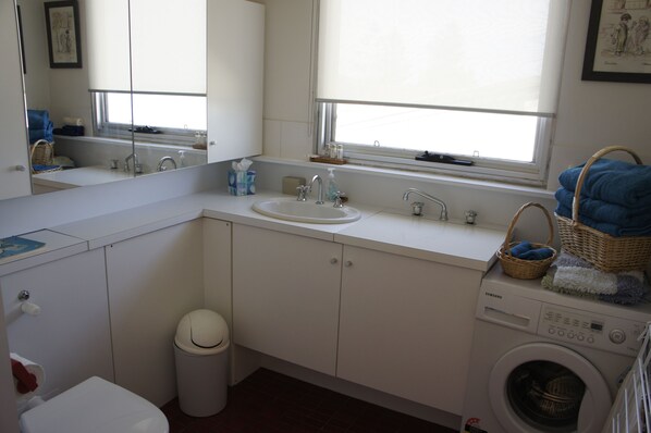 Un baño pequeño y luminoso con lavamanos blanco, inodoro, lavadora y ventana que proporciona luz natural.