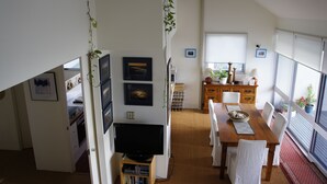 Un coin repas avec une table en bois, des chaises et un téléviseur. Des tableaux encadrés ornent le mur, il y a une fenêtre avec des stores et une plante suspendue au plafond.