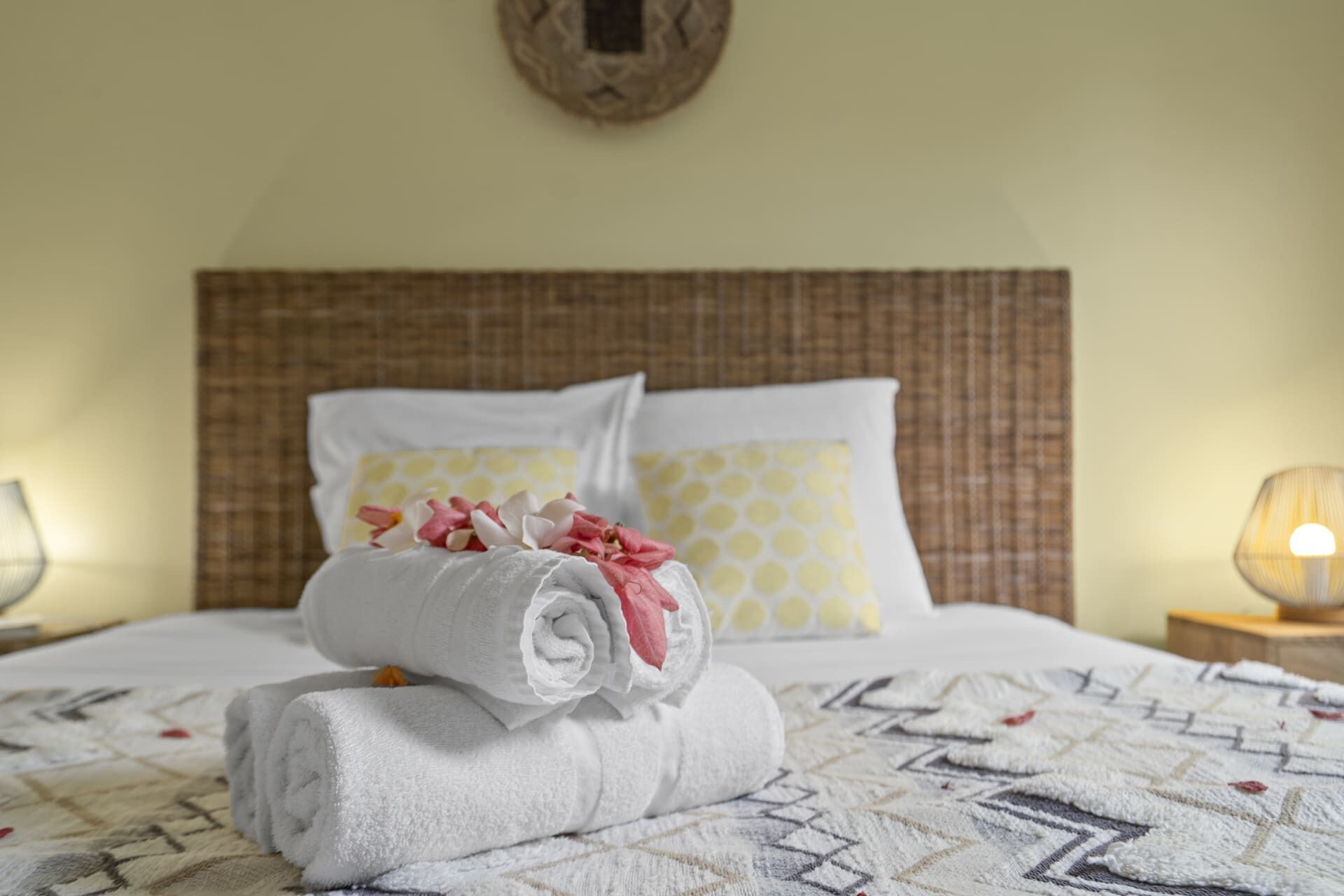 A neatly made bed with a rolled white towel and pink flowers, two patterned pillows, and a wall clock.