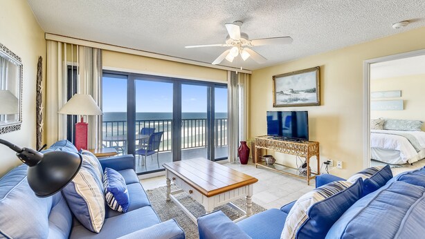 A spacious living area with a large sofa, a coffee table, and a television. There is a balcony with a view of the ocean.