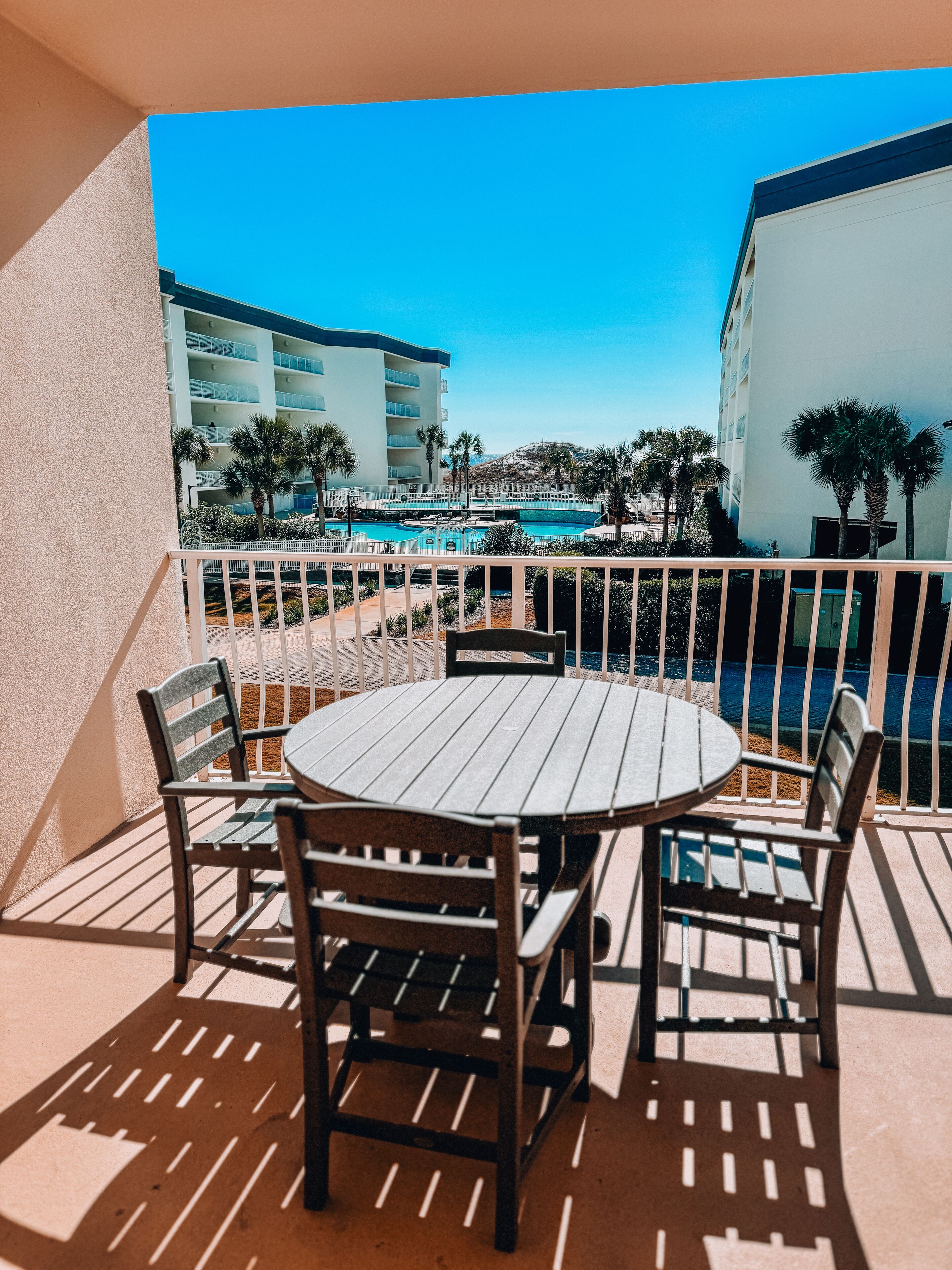 Een balkon met een ronde houten tafel en vier stoelen, met uitzicht op een zwembad en een gebouw met meerdere balkons.