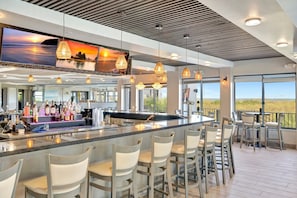 A modern bar area with a long counter, multiple bar stools, and a large TV screen displaying scenic views.