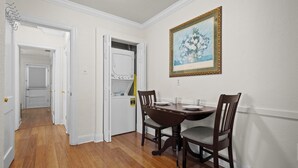 Un coin repas avec une table et des chaises en bois, un mur blanc orné d’un tableau encadré et un couloir menant à une autre pièce.