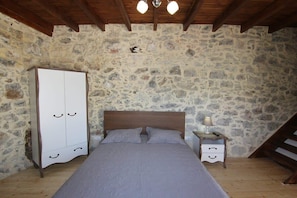A bedroom with a stone wall, a wooden ceiling, a bed with a grey bedspread, a white wardrobe, and a nightstand with a lamp.