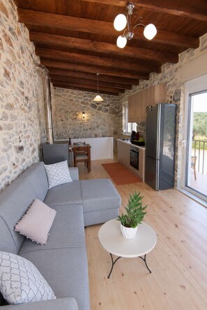 A cozy interior with a stone wall, a grey sofa, a small table with a plant, and a kitchen area with a refrigerator and stove.