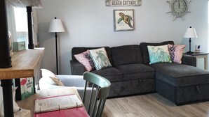 Uma sala de estar aconchegante com um sofá cinza escuro, uma mesa de centro de madeira e uma poltrona verde-água. Um relógio de parede e uma obra de arte emoldurada estão à vista.