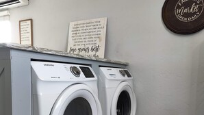 Uma área de lavanderia com duas máquinas de lavar Samsung, piso de madeira e uma parede com uma frase motivacional.