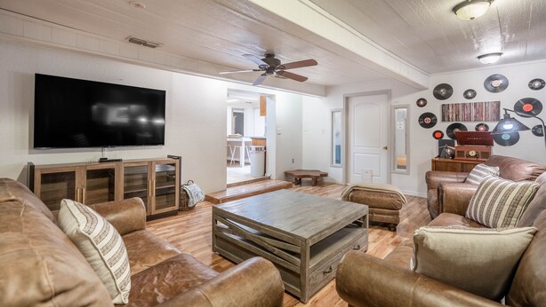 A living room with a flat-screen TV, wooden coffee table, leather sofas, and a ceiling fan.