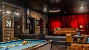 A billiards table, a sofa, and a red curtain in a room with brick walls and a tufted leather couch.