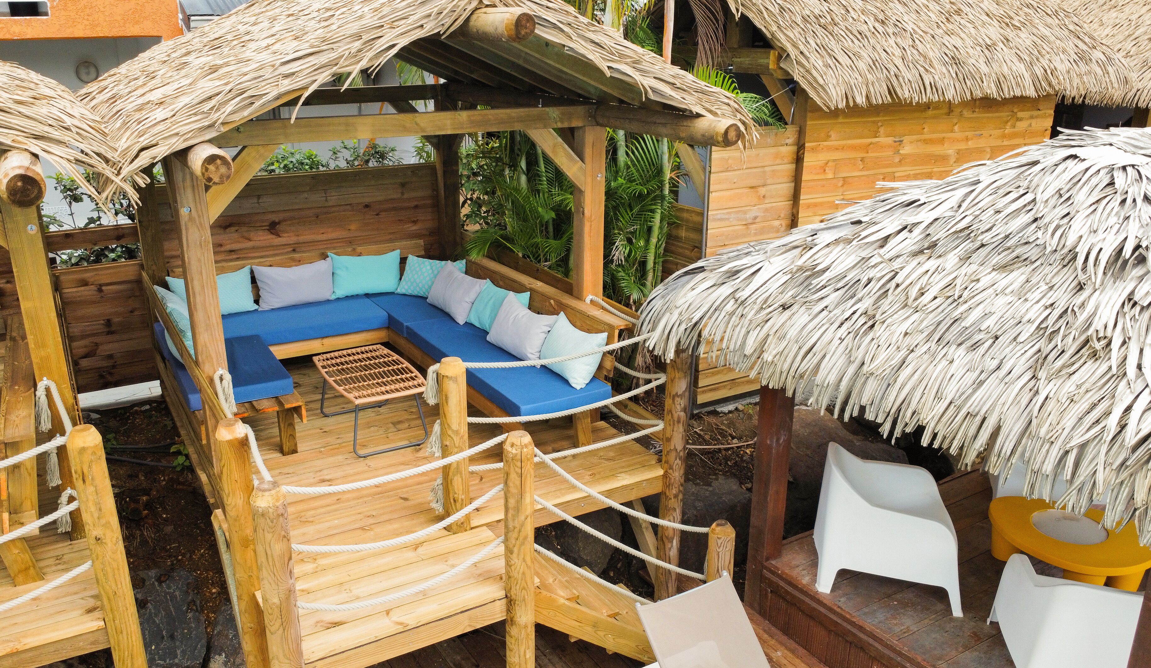 Une structure en bois surmontée d’un toit de chaume, dotée d’un espace salon avec des coussins bleus et d’une terrasse en bois avec des garde-corps en corde.