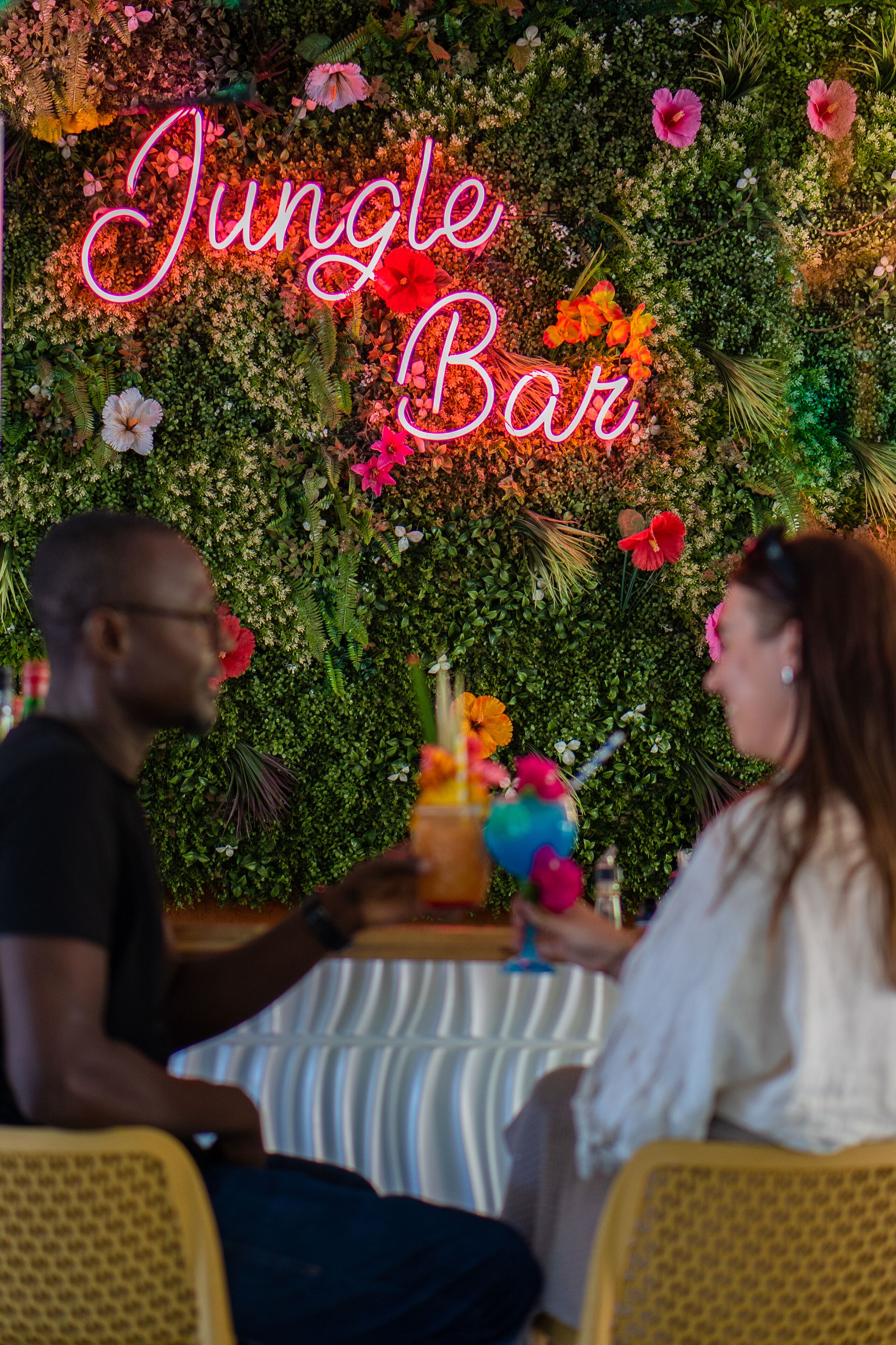 A couple seated at a table with a neon sign reading "Jungle Bar" in the background.