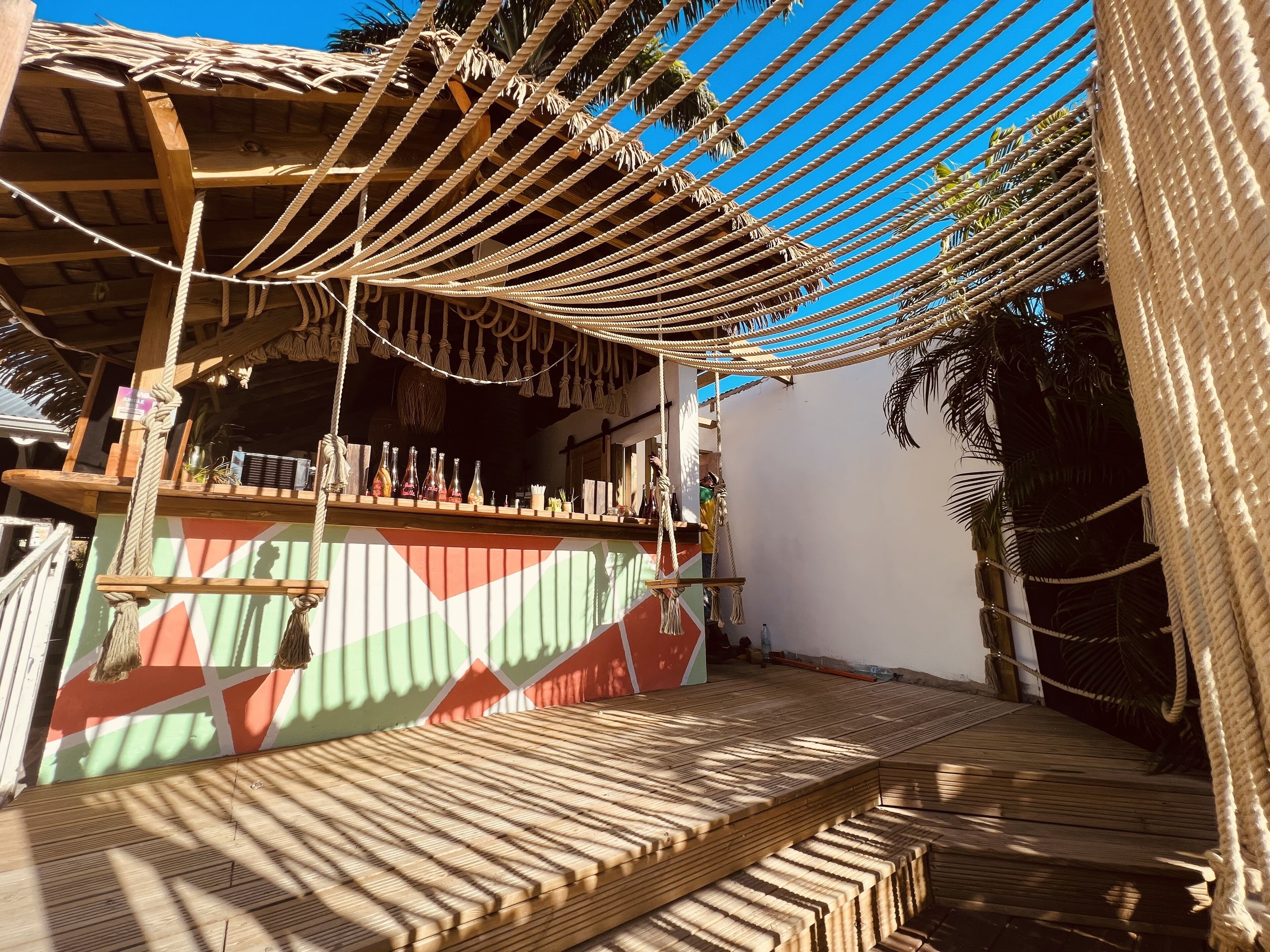 Una terraza de madera con techo de paja y cuerdas colgantes, que conduce a un área de bar con botellas y una pared colorida.