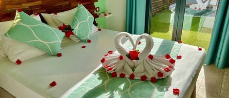 A neatly made bed with rose petals and a swan-shaped towel arrangement, a wooden headboard, and a view of a garden through a glass door.