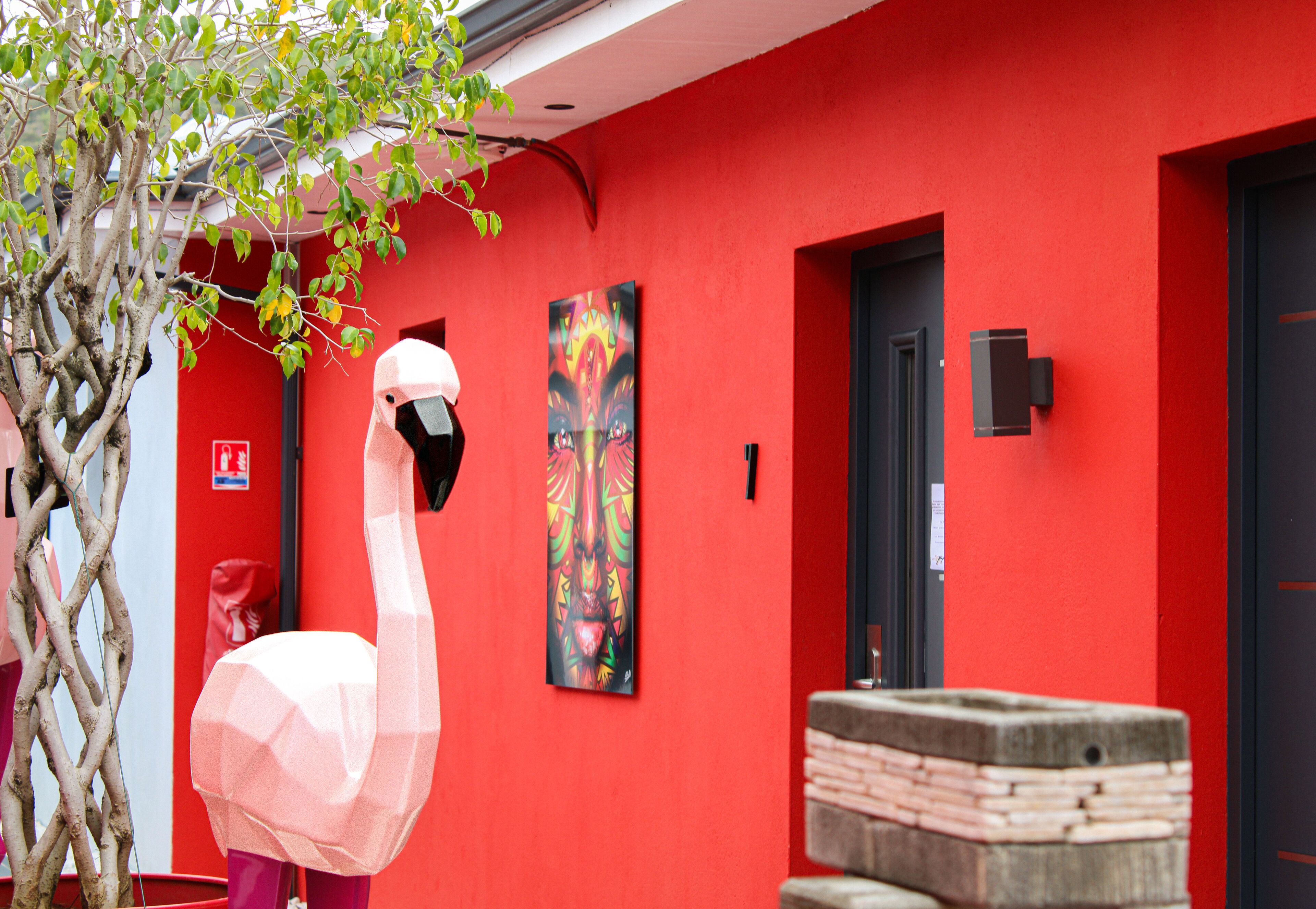 Un bâtiment rouge avec une grande sculpture de flamant rose rose devant, une porte et un panneau indiquant la sortie de secours.
