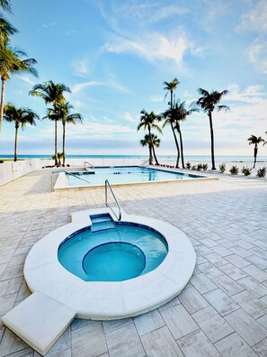 A swimming pool with a hot tub, surrounded by palm trees and a view of the ocean. - Top Floor Beachfront & Sunset Views! Walk to Everything! Smart TVs + Workspace! (Fort Myers Beach)