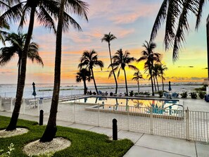 A pool area with palm trees, a fence, and a view of the beach during sunset. - Top Floor Beachfront & Sunset Views! Walk to Everything! Smart TVs + Workspace! (Fort Myers Beach)