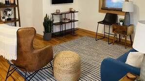 A living room with a ceiling fan, a flat-screen TV, a bookshelf, a coffee table, a blue sofa, and a patterned rug. - Premier Downstairs Flat in Perfect Down Town Location  (Ashland)