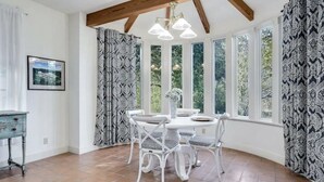 Ein Essbereich mit einem runden Tisch und weißen Stühlen, großen Fenstern mit gemusterten Vorhängen sowie einem Holzbalken an der Decke.