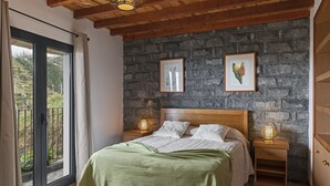 A bedroom with a stone wall, a bed with a green blanket, two bedside lamps, and a view through a glass door.