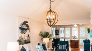 Ein geräumiges Wohnzimmer mit Couch, Sessel und einem Holztisch. Eine Deckenleuchte hängt von der Decke, und an der Wand hängt ein Gemälde eines Vogels.