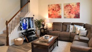 A living room with a brown leather sofa, a flat-screen TV, a ceiling fan, and floral wall art. - Spacious Luxurious 6 Bedroom 5 Bath Villa Sleeps 12 Pool, Jacuzzi, near Disney (Kissimmee)