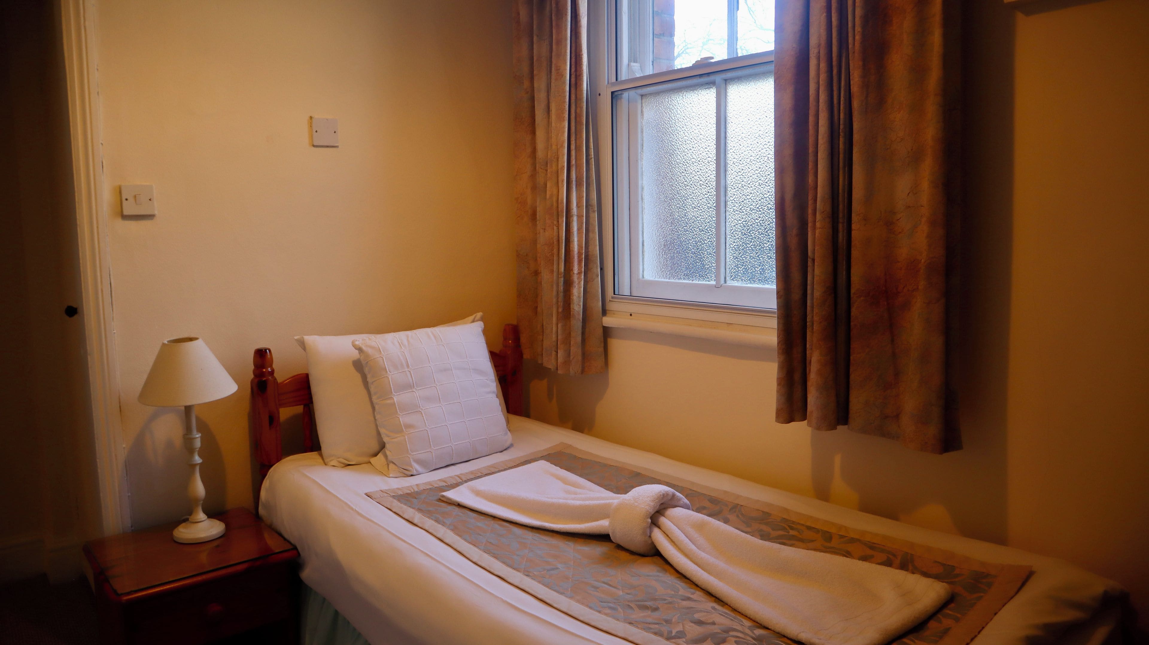 A single bed with a white duvet and a patterned blanket, a bedside table with a lamp, and a window with curtains.