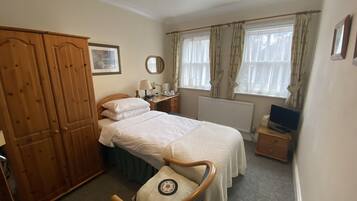 A bedroom with a bed, wooden wardrobe, bedside table with lamp, mirror, and a window with curtains.