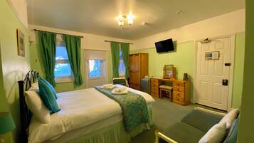A bedroom with a bed, a desk, a chair, a bedside table, a TV, and a door.