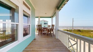 A wooden deck with a table and chairs, overlooking a beach and a building in the distance. - Salty Waves: 3  BR, 2  BA House in Port Bolivar, Sleeps 12 (Bolivar Peninsula)