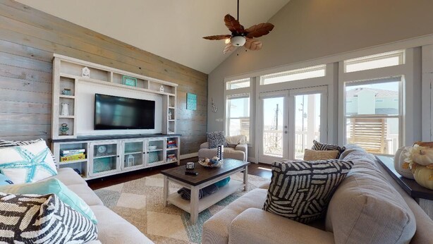 A living room with a flat-screen TV, a ceiling fan, and a large window. - Salty Waves: 3  BR, 2  BA House in Port Bolivar, Sleeps 12 (Bolivar Peninsula)