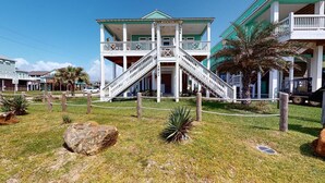 A two-story building with a central staircase, a covered porch, and a fenced front yard with palm trees and rocks. - Salty Waves: 3  BR, 2  BA House in Port Bolivar, Sleeps 12 (Bolivar Peninsula)