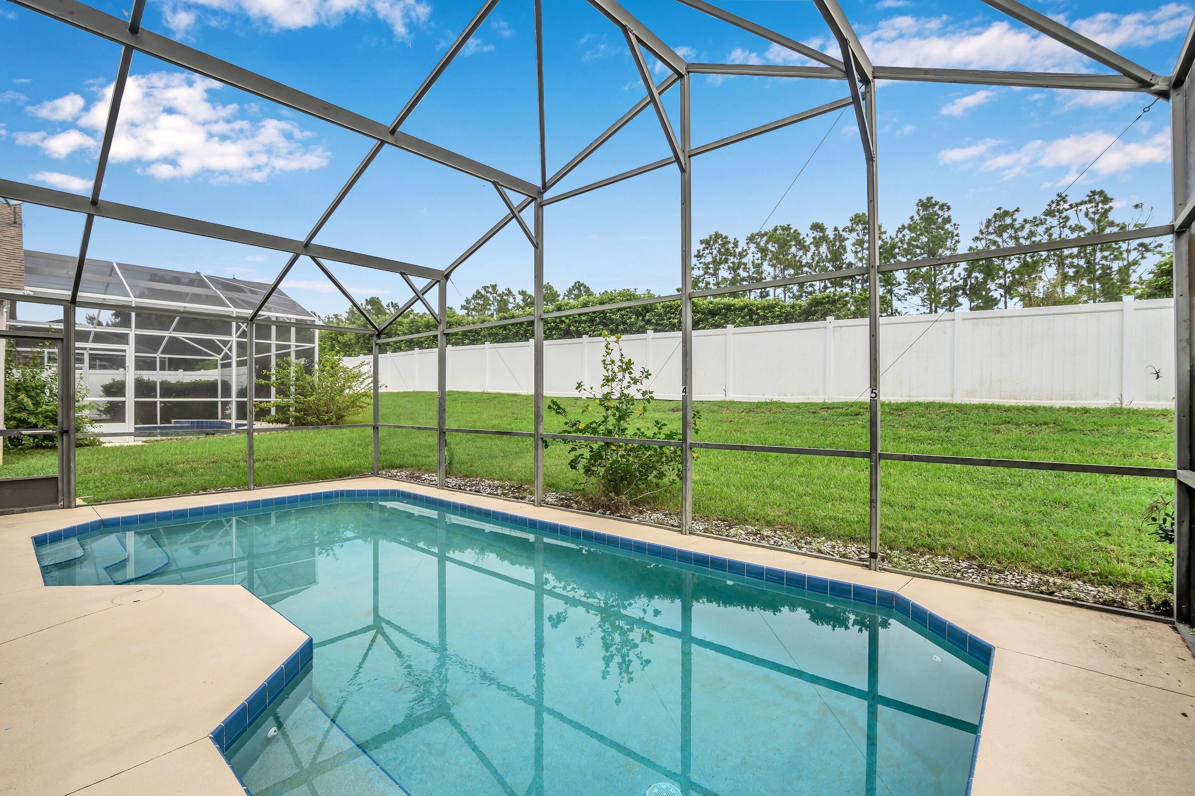 A swimming pool with a clear blue water, surrounded by a metal frame structure, and a grassy yard with a white fence in the background.