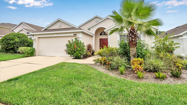 A suburban home with a garage, a red door, and a well-maintained lawn with various plants and a palm tree. - No Rear Neighbors, Near Disney Spacious Home With CDC Standards #3hr836 (Davenport)