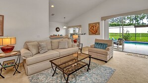 A spacious living room with a beige sofa, a glass coffee table, and a patterned rug. There is a dining area with a patterned chair and a table. A large window offers a view of a swimming pool and greenery. - No Rear Neighbors, Near Disney Spacious Home With CDC Standards #3hr836 (Davenport)