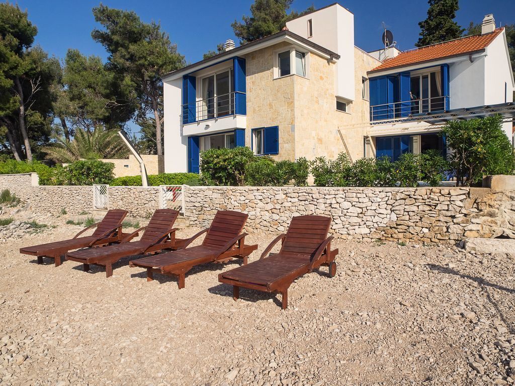 A modern two-story house with a stone wall and a pool area with wooden lounge chairs.