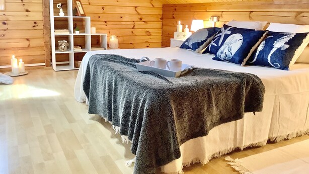 A cozy bedroom with a wooden ceiling, a bed with a dark blanket, bedside lamps, and a shelf with decorative items.