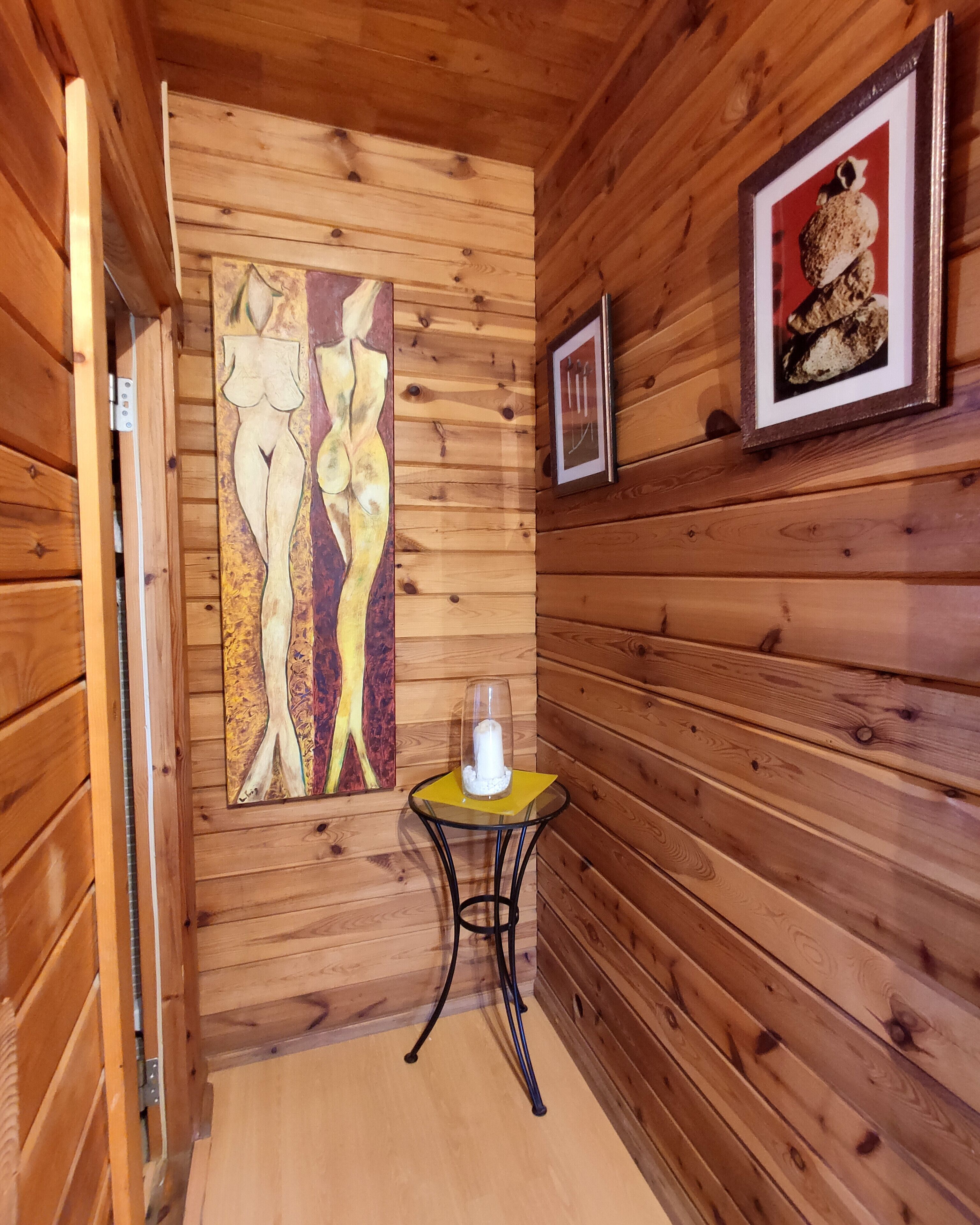 Interior de una cabaña de madera, con un muro de madera, un cuadro enmarcado, una mesita con una vela y suelo de madera.